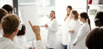 Eine Lehrveranstaltung im HSHL-Labor mit einer Dozentin und mehreren Studierenden im Kittel, wovon ein Student aufzeigt.